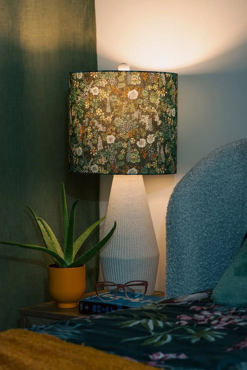 Botanic drum lampshade in Liberty Ciara floral cotton, glowing on a wooden side table in a cozy bedroom