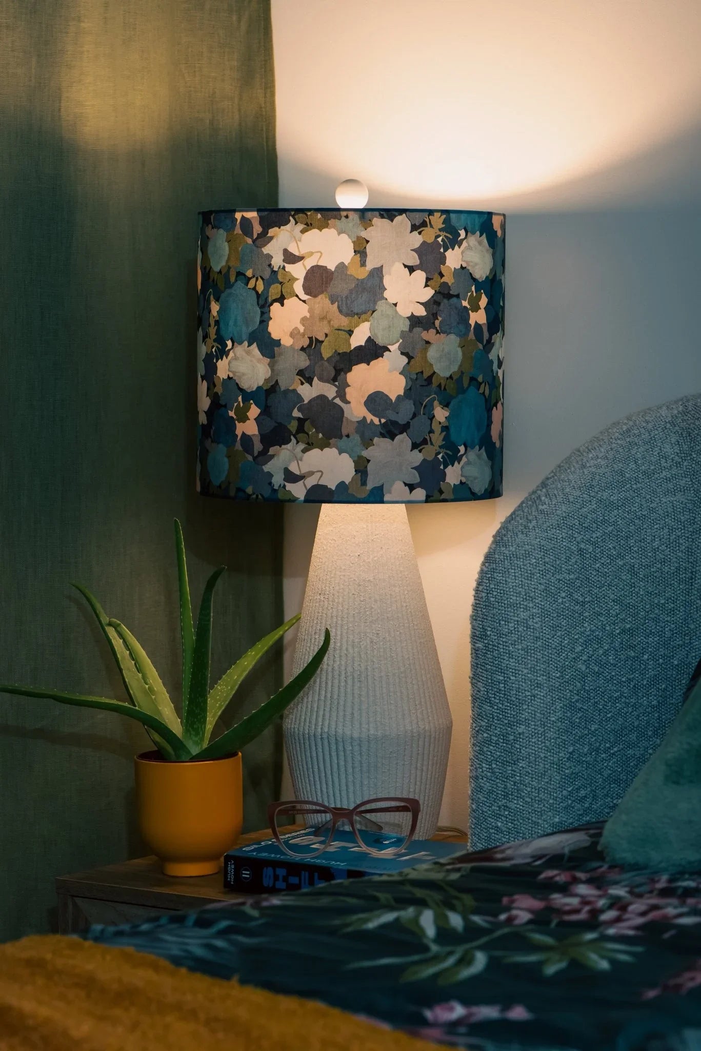 Something Blue drum lampshade in Liberty Little Merton cotton, glowing on a wooden bedside table in a cozy bedroom