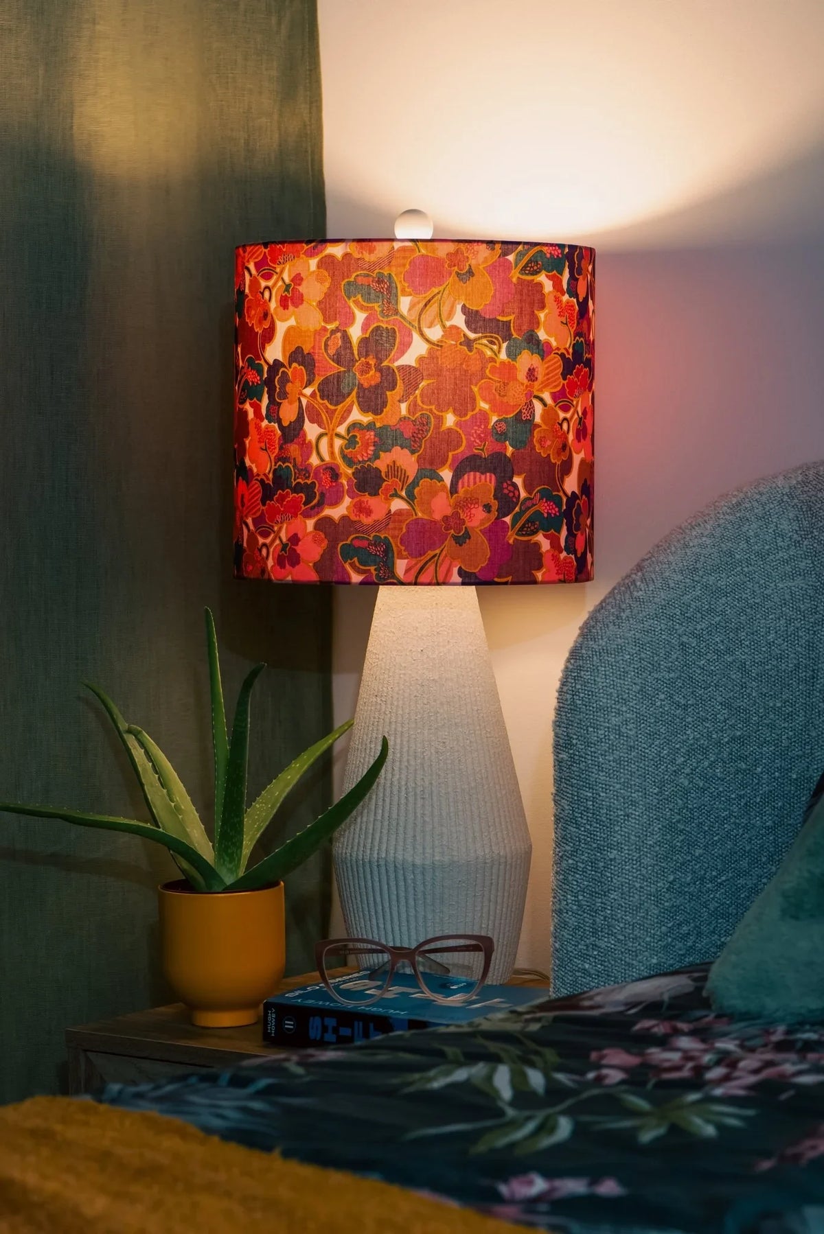 Sunflare drum lampshade in Liberty Prospect Flower cotton, casting a golden glow on a bedside table in a warm bedroom.